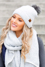 Woman wearing a white knit beanie with a pom-pom and a gray scarf against a neutral background