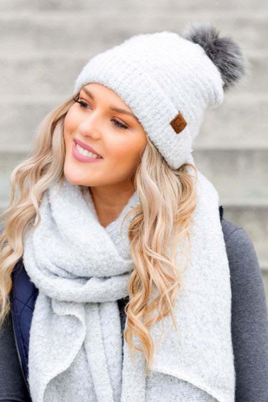 Woman wearing a white knit beanie with a pom-pom and a gray scarf against a neutral background