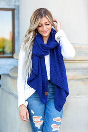 Woman wearing a blue scarf and white sweater with ripped jeans, standing against a light-colored wall.
