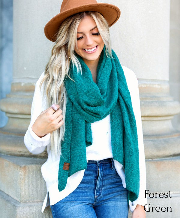 Woman wearing a forest green scarf, white shirt, blue jeans, and brown hat against a stone wall.
