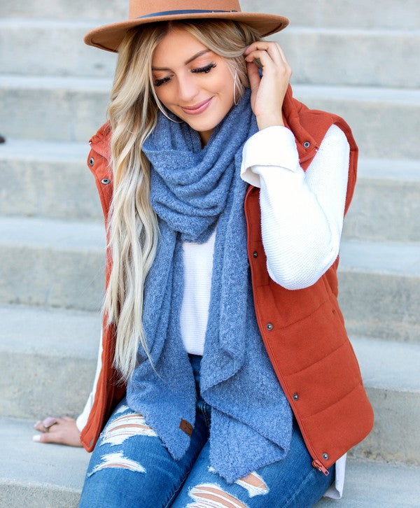 Woman wearing a brown vest, blue scarf, and jeans sitting on steps.