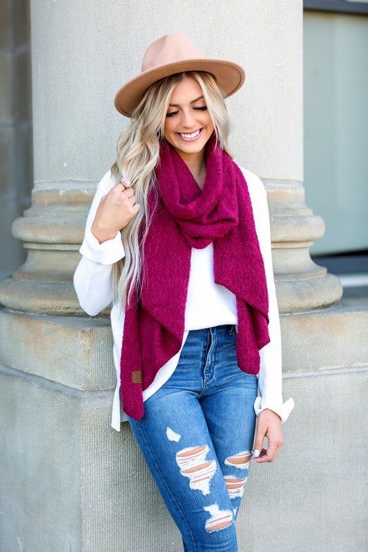 Woman wearing a pink scarf, white sweater, and blue jeans standing against a stone wall.