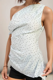 Woman wearing the celeste light blue one-shoulder top with black polka dots against a beige background