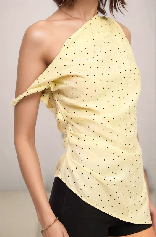a close up of the celeste yellow polka dot one-shoulder top worn by a woman against a neutral background