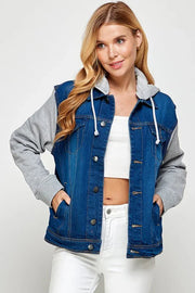 Woman wearing a dark blue denim jacket with gray sleeves over a white crop top has her hands in her pockets and looking to the side.