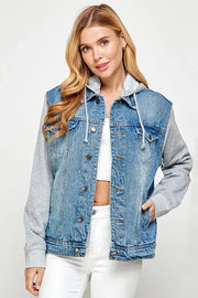 Woman wearing a denim jacket with a gray hoodie over a white crop top and white jeans looking at the camera with a hand in her pocket. 