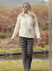 Woman wearing a cream sweater and plaid pants standing outdoors with a scenic background.