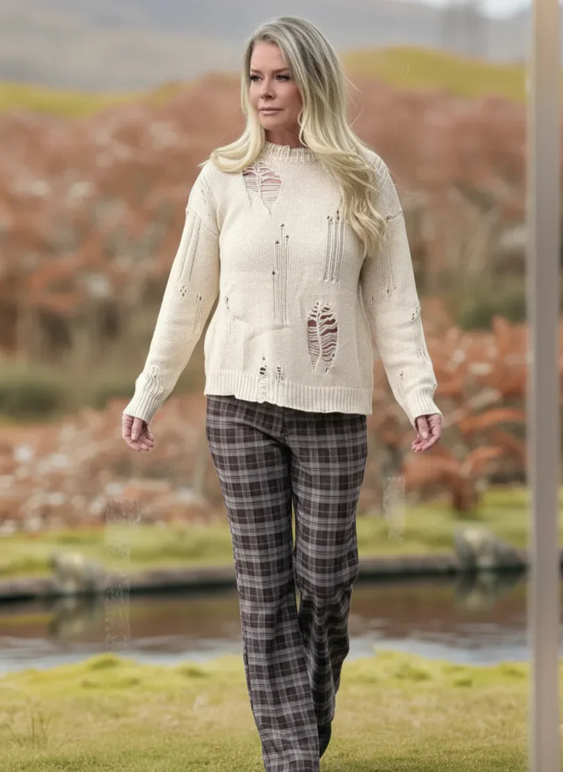 Woman wearing a cream sweater and plaid pants standing outdoors with a scenic background.