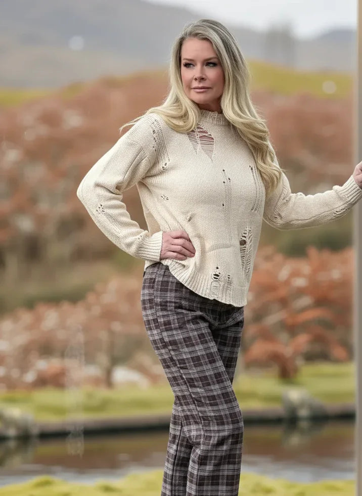 Woman in a cream sweater and plaid pants standing by a wooden post with a blurred natural background