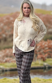 Woman wearing a distressed white sweater and plaid pants standing outdoors with a blurred natural background
