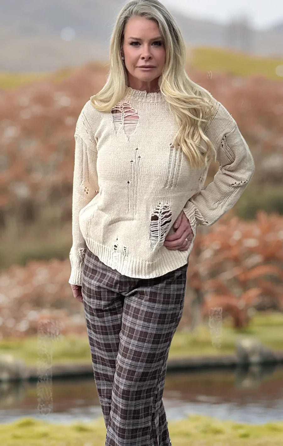Woman wearing a distressed white sweater and plaid pants standing outdoors with a blurred natural background