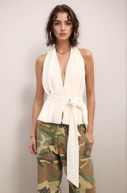 Woman wearing the elysian white sleeveless halter top and camouflage pants against a plain background