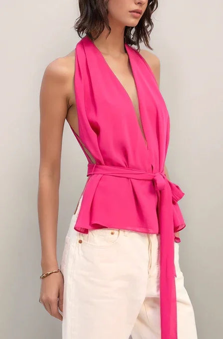 Woman wearing the elysian bright pink sleeveless top with a plunging neckline and white pants against a plain background.