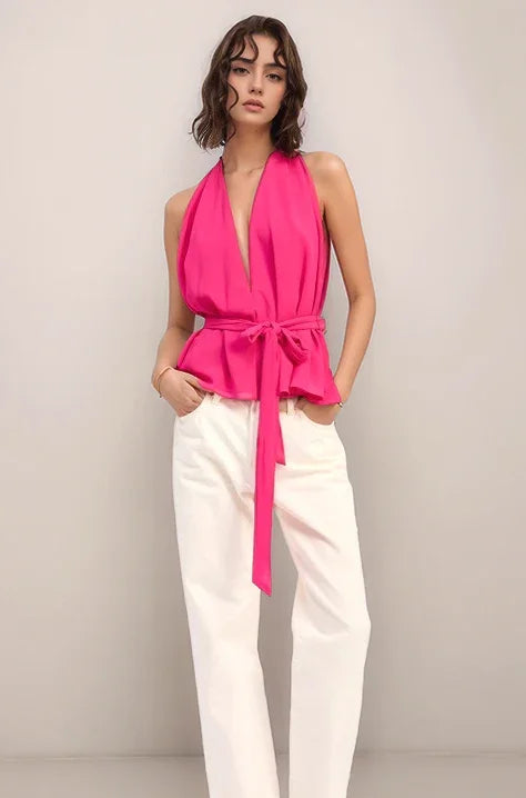Woman wearing the elysian hot pink halter top  with a sash tie and white pants against a plain background