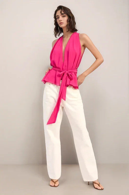 Woman wearing the hot pink elysian sleeveless halter top and white pants against a plain background