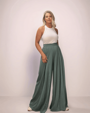 a blonde woman wearing a pair of sage green wide leg pants and a white tank top. The pants are long and wide leg. They also have pleats in the front.