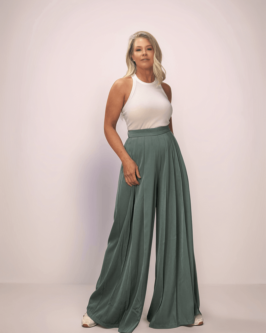 a blonde woman wearing a pair of sage green wide leg pants and a white tank top. The pants are long and wide leg. They also have pleats in the front.