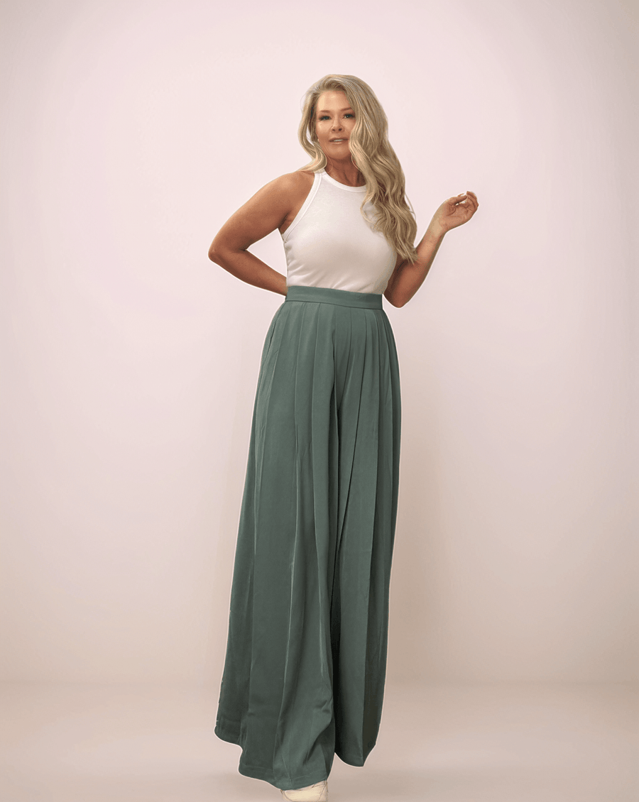 a blonde woman wearing a pair of sage green wide leg pants and a white tank top. The pants are long and wide leg. They also have pleats in the front.