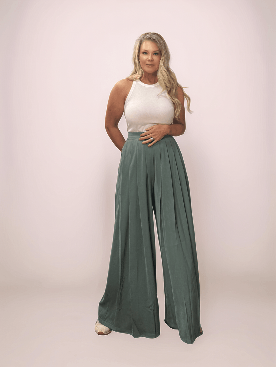 a blonde woman wearing a pair of sage green wide leg pants and a white tank top. The pants are long and wide leg. They also have pleats in the front.