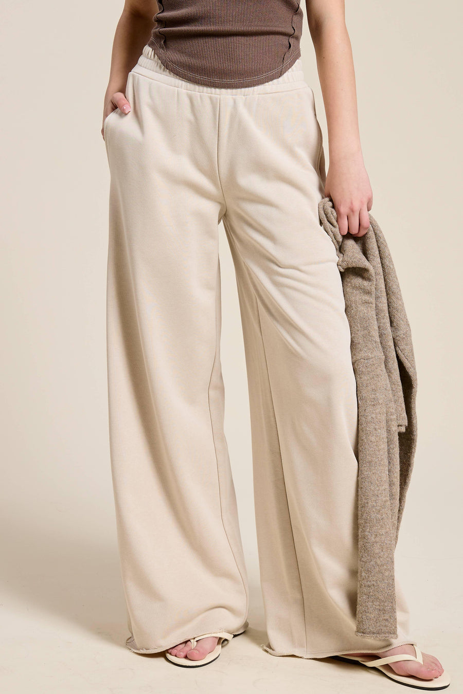 Person wearing beige wide-leg pants holding a brown jacket against a plain background