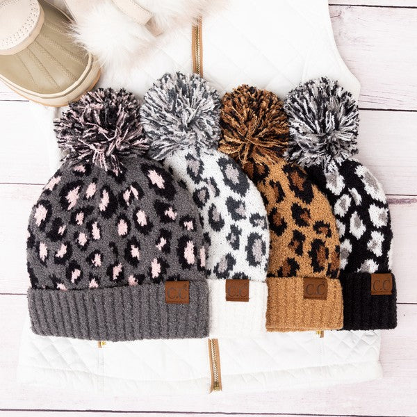 Four leopard print beanies with pom-poms on a white surface
