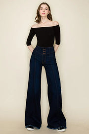 Woman wearing a black off-shoulder top and dark blue flared jeans on a beige background