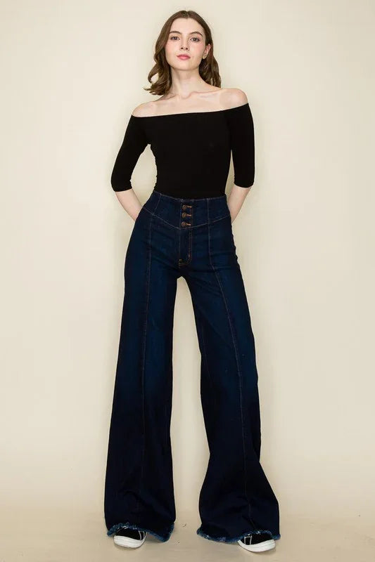 Woman wearing a black off-shoulder top and dark blue flared jeans on a beige background