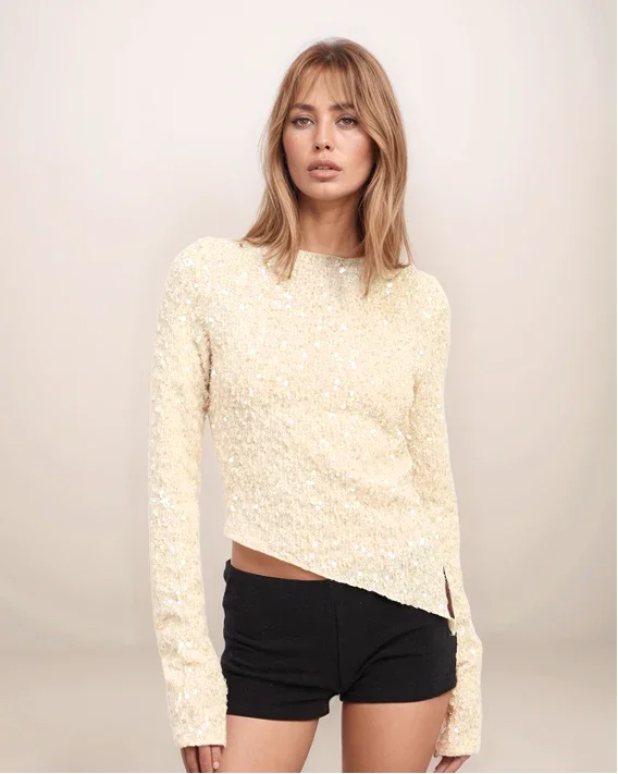 Woman wearing a cream-colored long-sleeve sequin top and black shorts against a plain background