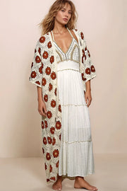 A woman wearing the Dahlia Floral Crochet Cardigan in cream with terracotta flower motifs layered over a white tiered maxi dress.