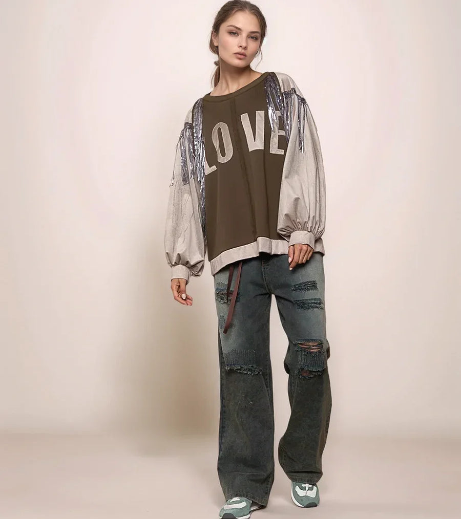 Woman wearing a brown 'LOVE' shirt with sheer sleeves and ripped jeans on a beige background