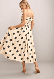 a blonde woman wearing a cream colored dress with black polka dots. It has a ruched bodice, spaghetti straps and a full skirt. The woman is holding the dress out to the side. 