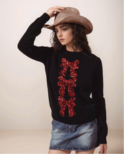 Woman wearing a black sweater with red sequin letters, denim skirt, and cowboy hat on a plain background