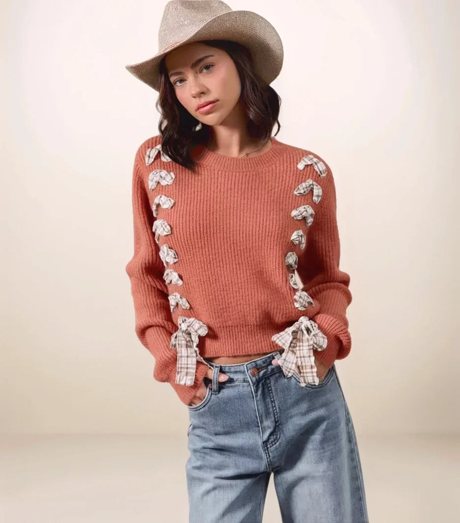 Woman wearing a rust-colored sweater with plaid accents, blue jeans, and a beige hat on a plain background