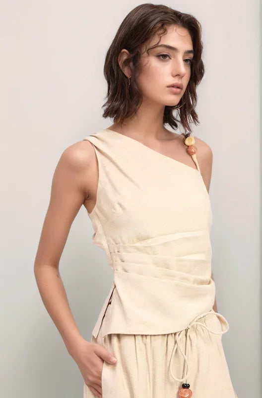 an angled view of the sand-colored Sonora Asymmetrical Top, featuring a sophisticated one-shoulder neckline and flattering ruched pleating along the side.