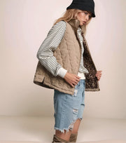 Woman wearing a beige quilted vest over a striped shirt with denim shorts on a plain background