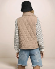 Person wearing a beige quilted vest over a striped shirt with denim shorts on a plain background