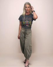 A woman wearing a dark grey Def Leppard band t-shirt, faded light gray relaxed-fit bubble pants with a drawstring, and black flip-flops.