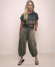 A woman wearing a dark grey Def Leppard band t-shirt, olive green relaxed-fit bubble pants with a drawstring, and black flip-flops with one hand in her pocket.